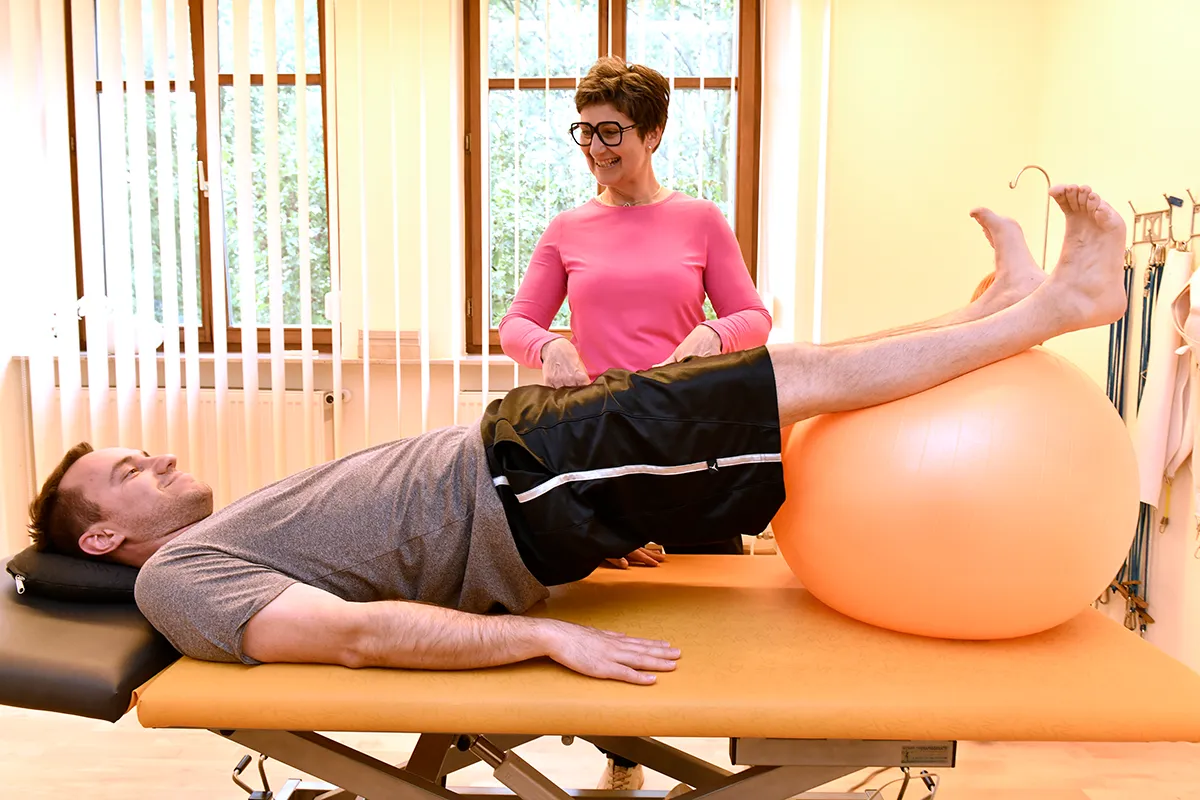 Ein Mann liegt lächelnd auf einer Liege, die Füße liegen oben auf einem Gymnastikball. Hinter ihm steht eine Physiotherapeutin in einem pinken Oberteil und schaut ihn lächelnd an.