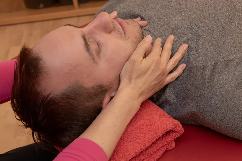 Ein Patient liegt mit geschlossenen Augen auf einer Liege mit einem Handtuch im Nacken. Eine Physiotherapeutin fasst ihn von links und rechts an den Hals.