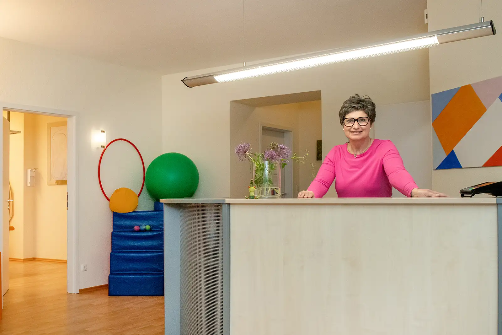 Empfangsbereich mit warmen Farben. Hinter dem Tresen steht eine freundlich lächelnde Frau mit Brille in einem pinken Oberteil.